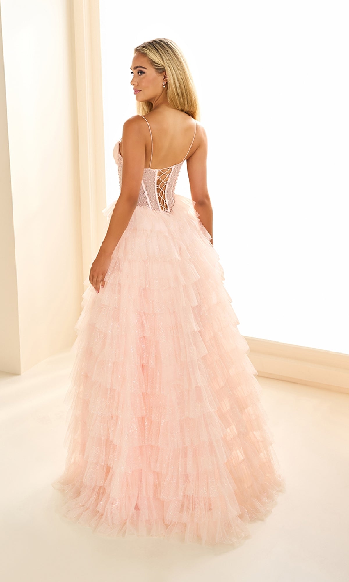 Woman in the Ellie Wilde EW36029 Ruffled Prom Ball Gown with a lace-up back and tiered ruffle tulle skirt, standing in a brightly lit, minimalist room, facing away from the camera.